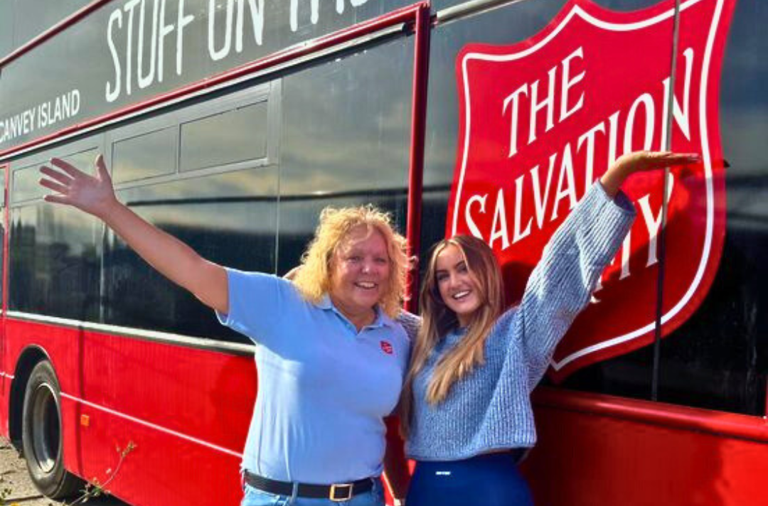 A photo shows two people standing with their arms raised outside a Salvation Army-branded double-decker bus.