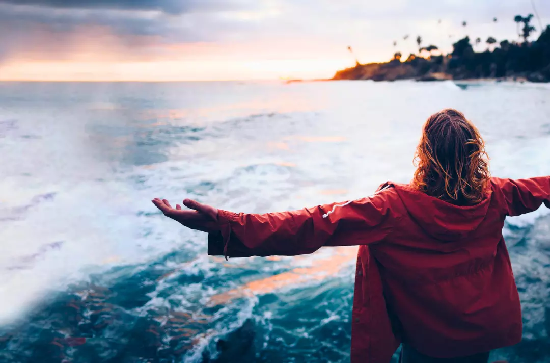 A photo shows someone standing with their arms outstretched in front of a roaring ocean. 
