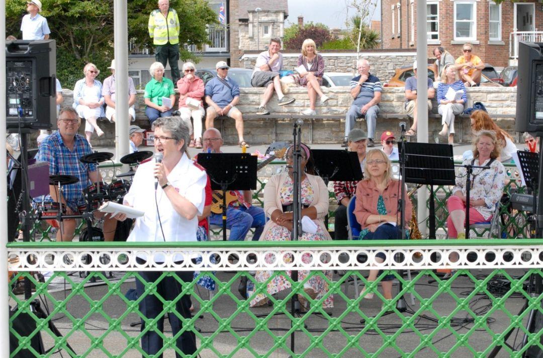 A photo of Major Katrina Greetham speaking through a microphone at an outside event in a bandstand