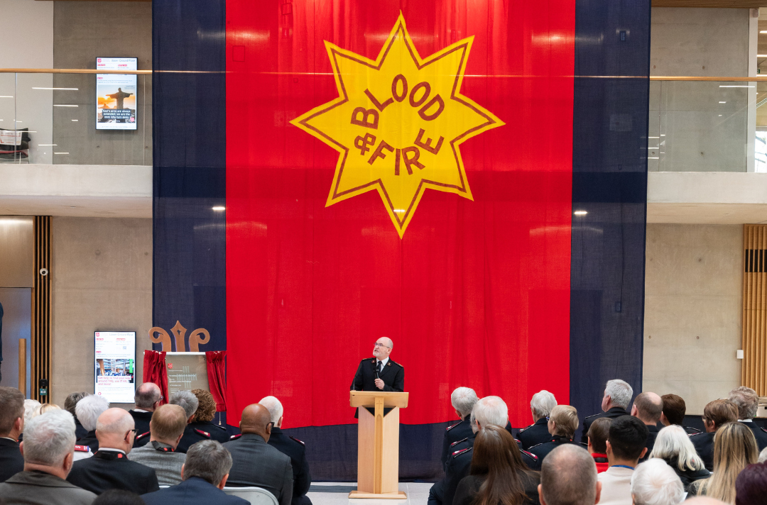 A photo shows General Lyndon Buckingham speaking at a podium in front of an enormous Salvation Army flag.