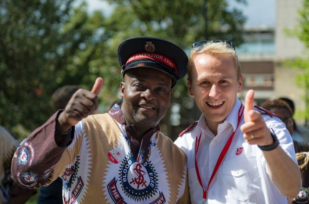 A photo of a black Salvationist wearing an African Salvation Army shirt and a white Salvationist wearing a white Salvation Army shirt. They are smiling at the camera with an arm around each other and their thumbs up 