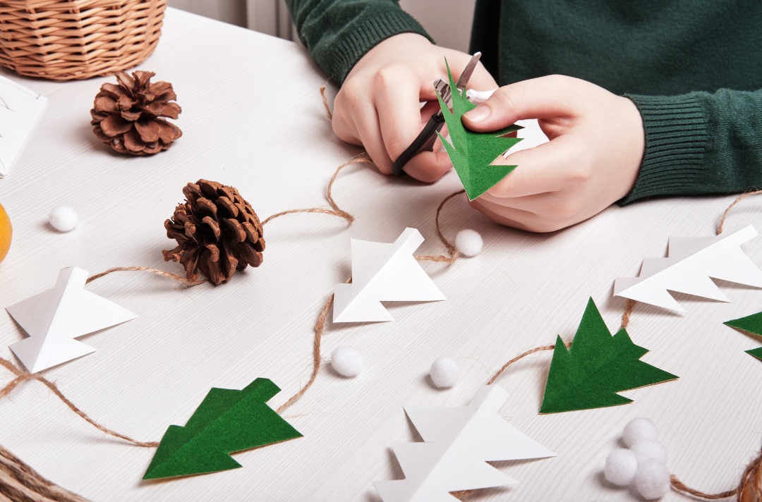 A photo shows someone constructing Christmas decorations out of paper, pinecones and string.