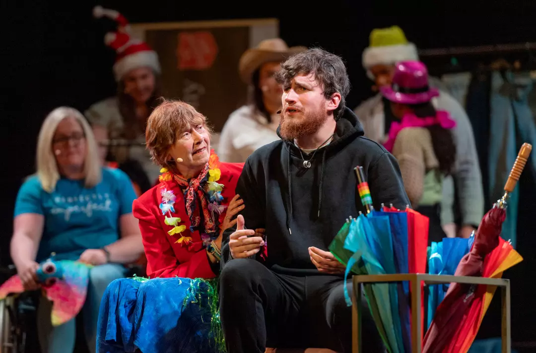 A photo of a scene from Belongings - an older woman has their hand on a mans arm as he sings