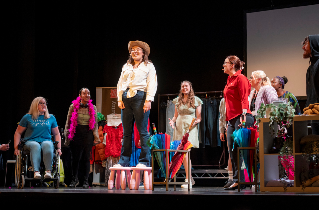 The cast of Belongings performing a song - a young woman is standing on a stool singing wearing a cowboy hat surrounding by other cast members
