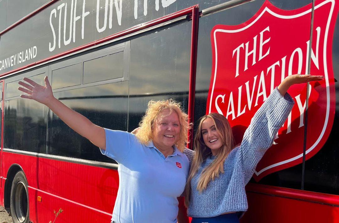 A photo of two women with their hands in the air standing in front of a red bus with the Salvation Army red shield on it 