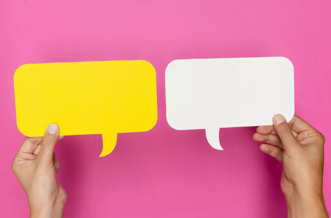 A photo shows two hands holding pieces of paper each cut into the shape of a speech bubble, one yellow, the other white, against a strong pink background.