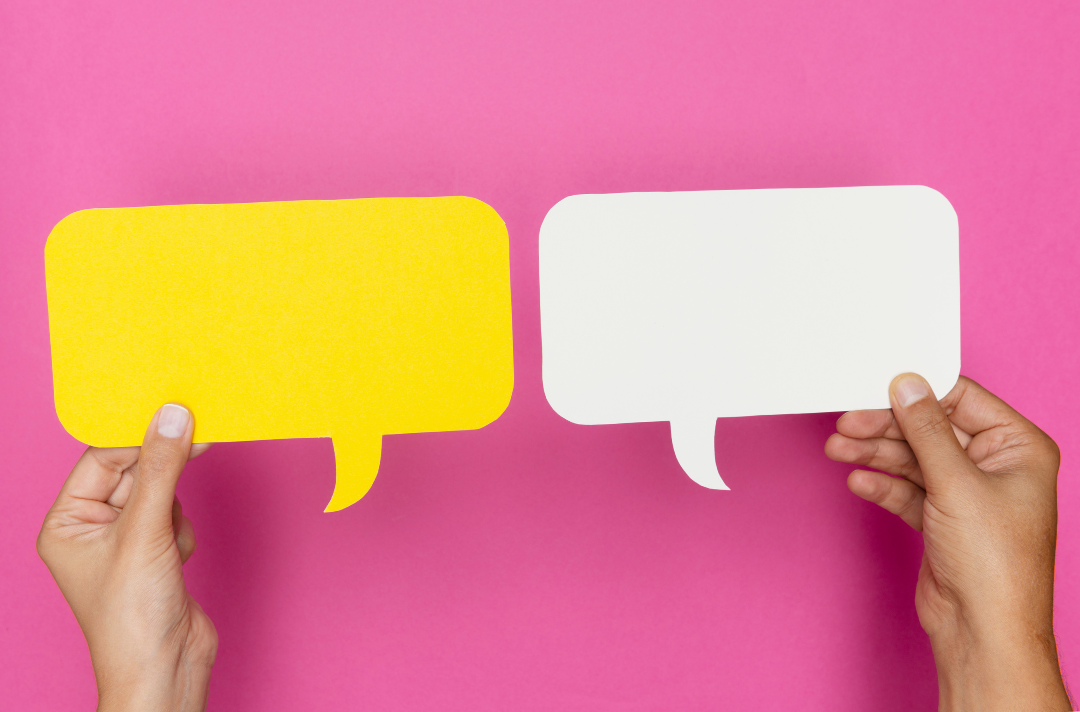 A photo shows two hands holding pieces of paper each cut into the shape of a speech bubble, one yellow, the other white, against a strong pink background.