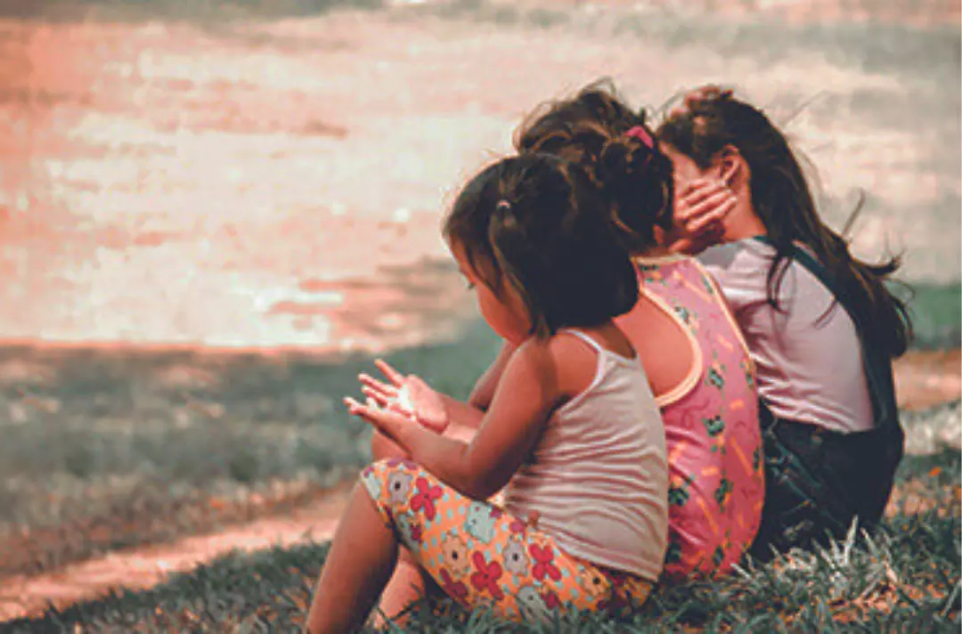 A photo shows three children sat on some grass by a large body of water.