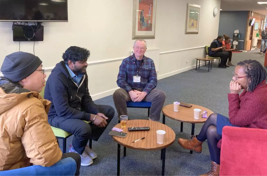A photo shows people from different backgrounds seated in comfy chairs around a table and having a conversation.