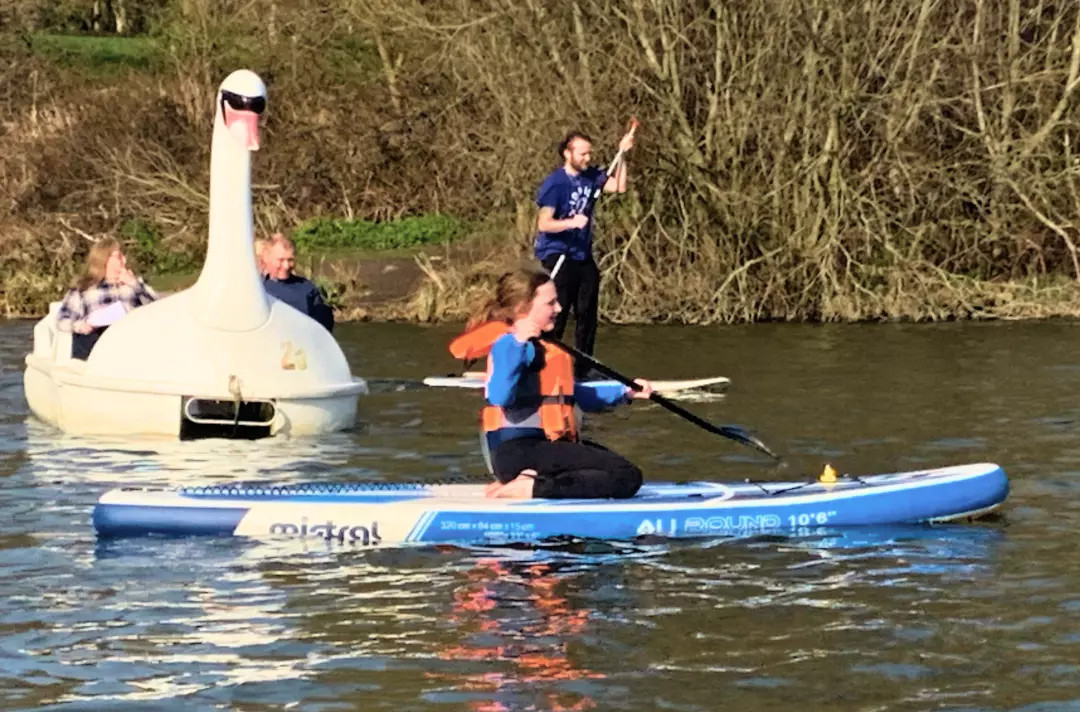 A photo shows someone paddle boarding. In the background some people pilot a pedalo swan.