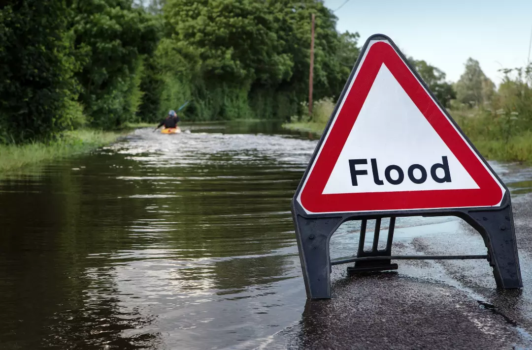 A photo shows a flooded country road. In the foreground, a triangular sign reads: Flood. In the distance, a person kayaks down the road.