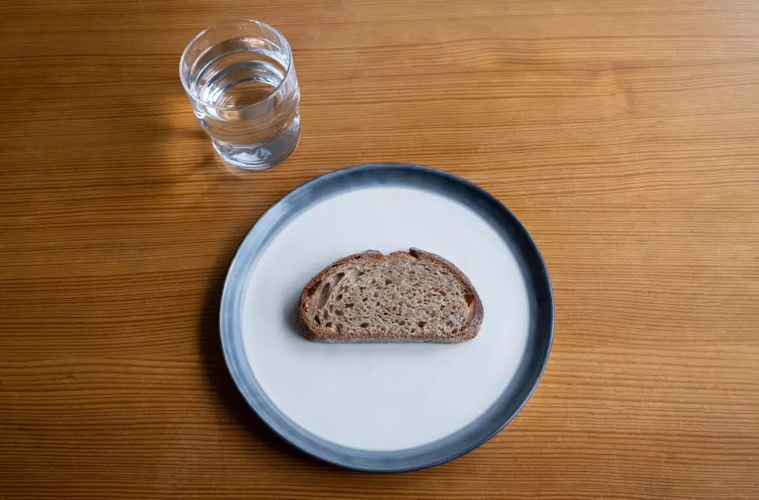A photo shows a simple table with small slice of bread on a plate and a glass of water.