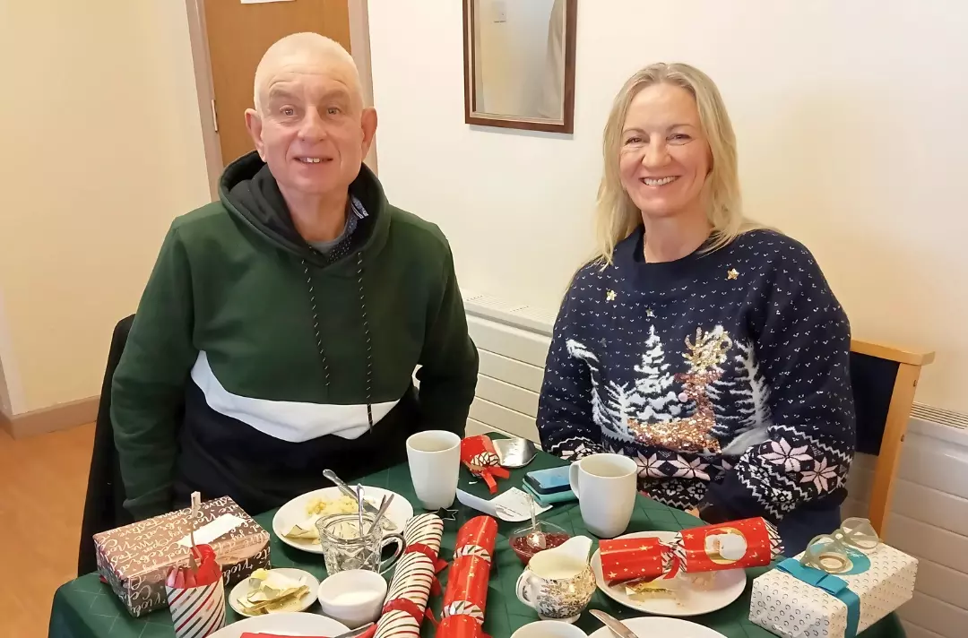 A photo shows two people smiling at the camera while sat at a table laid for Christmas dinner.