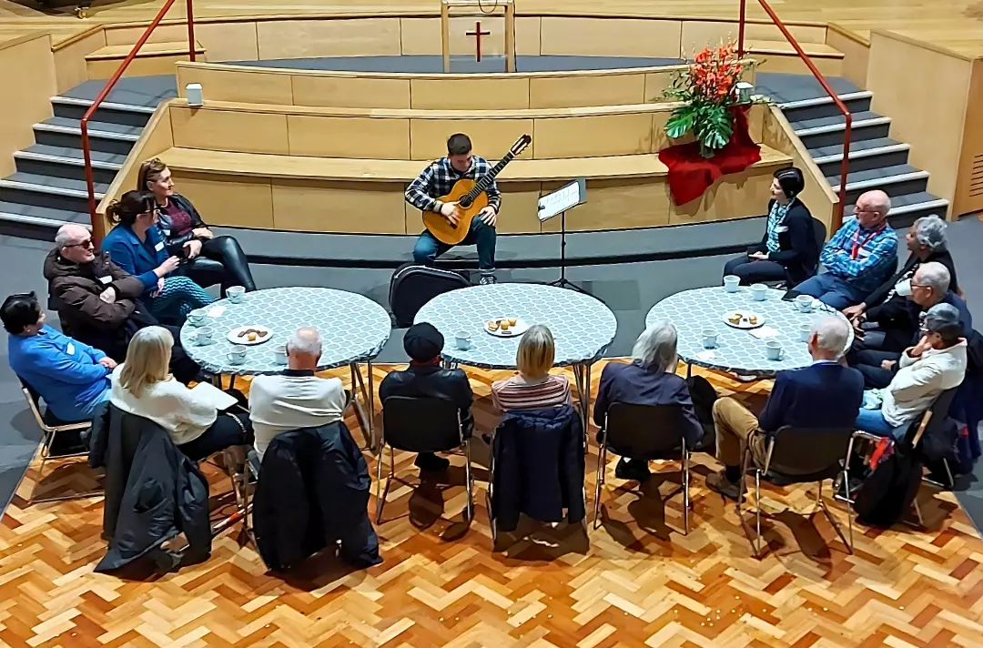 A photo shows people gathered around tables in a hall while someone at the front plays a guitar.