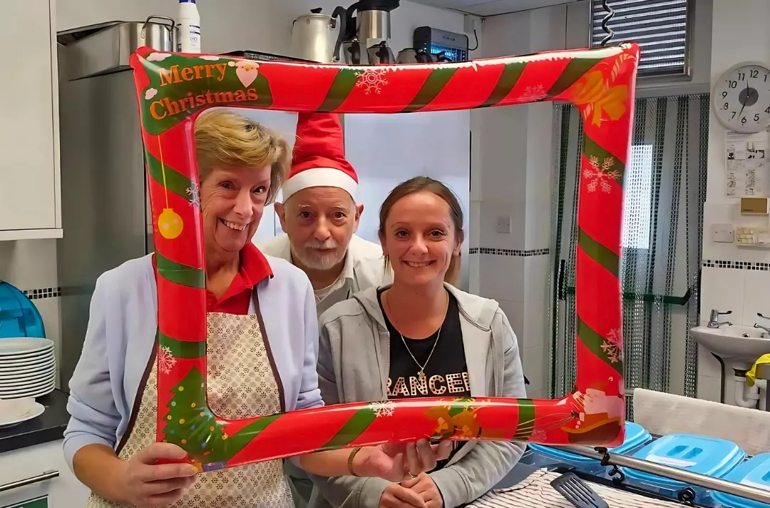 A photo shows three people in a Salvation Army kitchen posing behind an inflatable Christmas-themed picture frame.