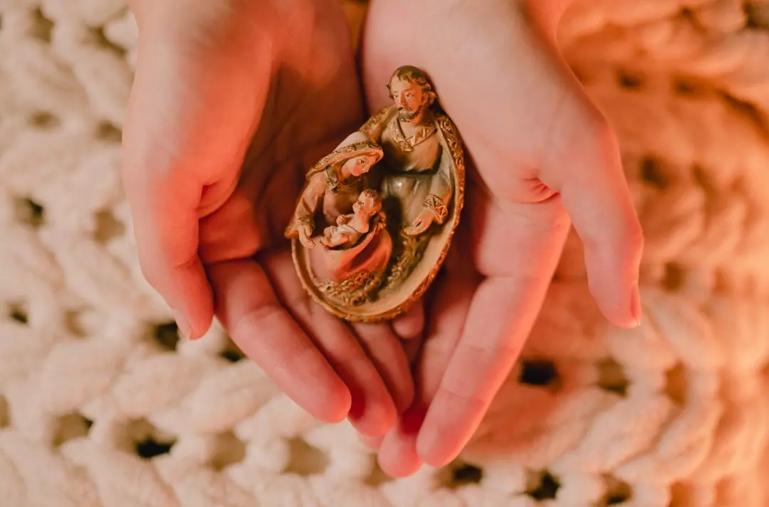A photo shows two hands gentle cupping a small wooden Nativity scene.