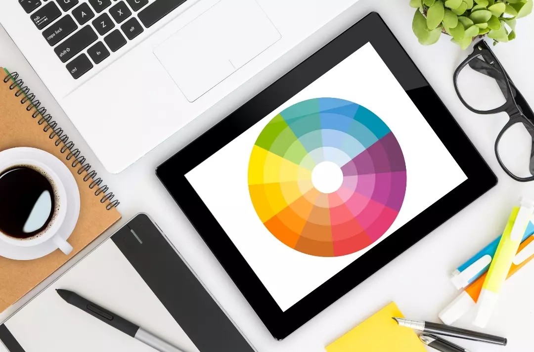 An image shows some of the tools used in graphic design, such as a laptop, tablet, pens, sticky notes and coffee.