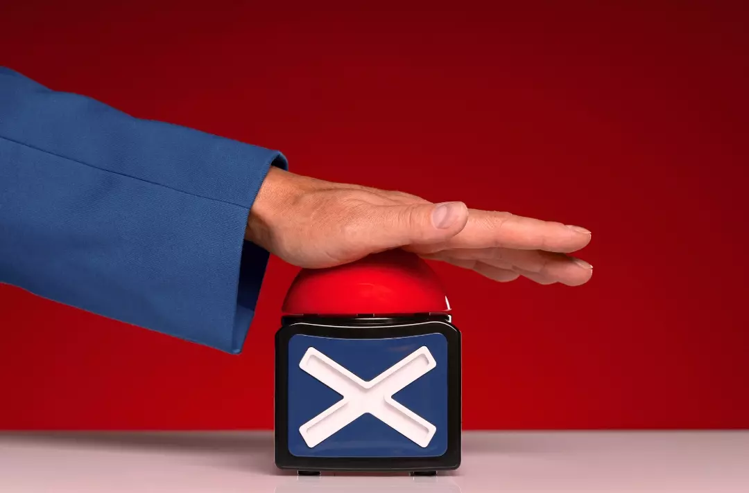 A photo shows a hand pressing a big, red buzzer with an X on it.