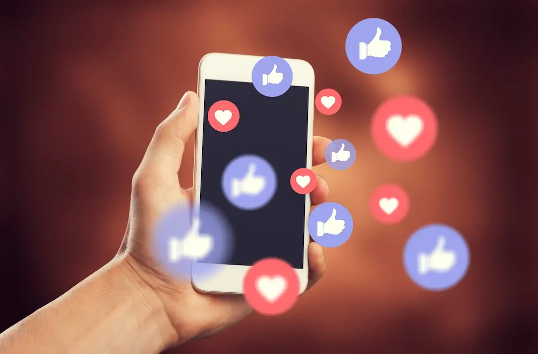A hand holds a phone with hearts and thumbs up reactions coming out of the screen