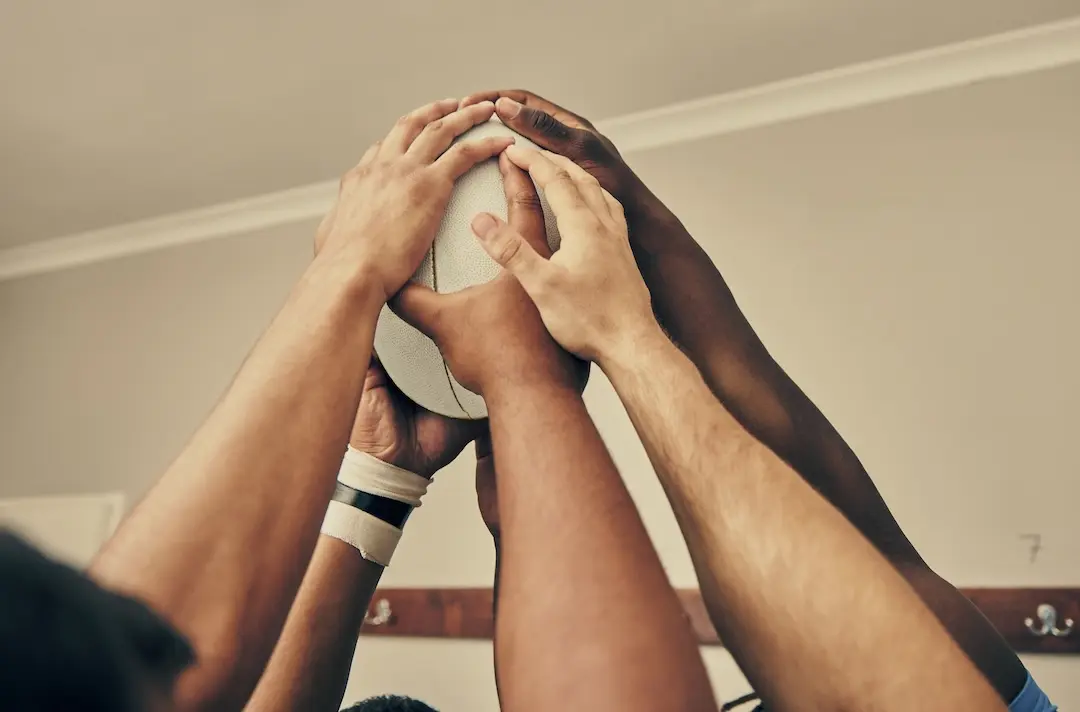 A mix of hands hold a rugby ball in a locker room.
