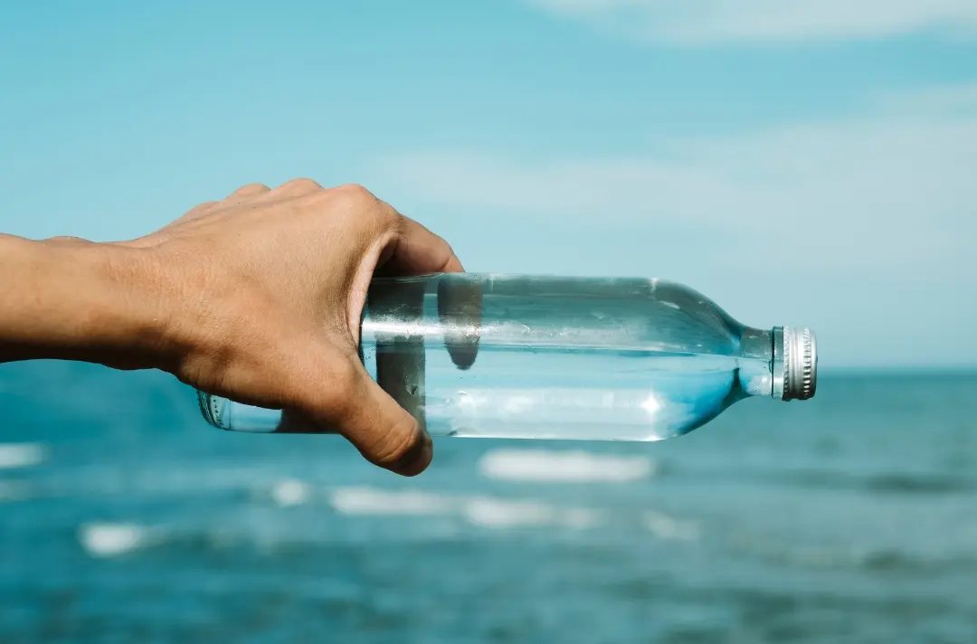 A photo shows someone holding a filled glass bottle in front of a vast ocean.