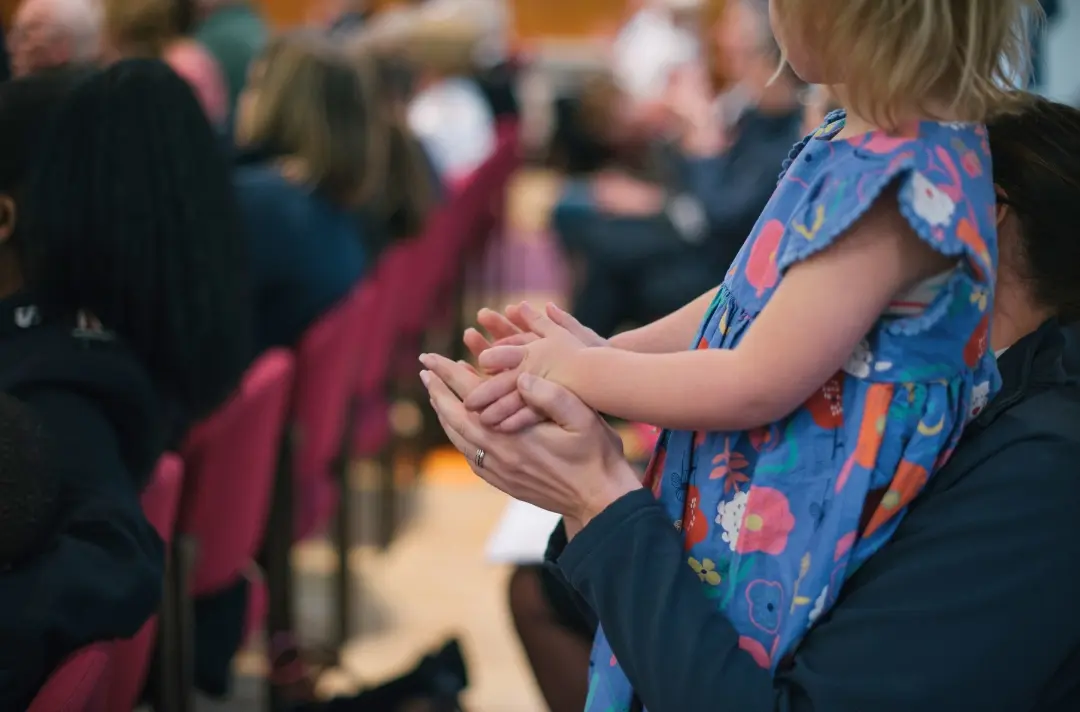 A photo shows a child standing on the lap of an adult during a Sunday morning meeting.