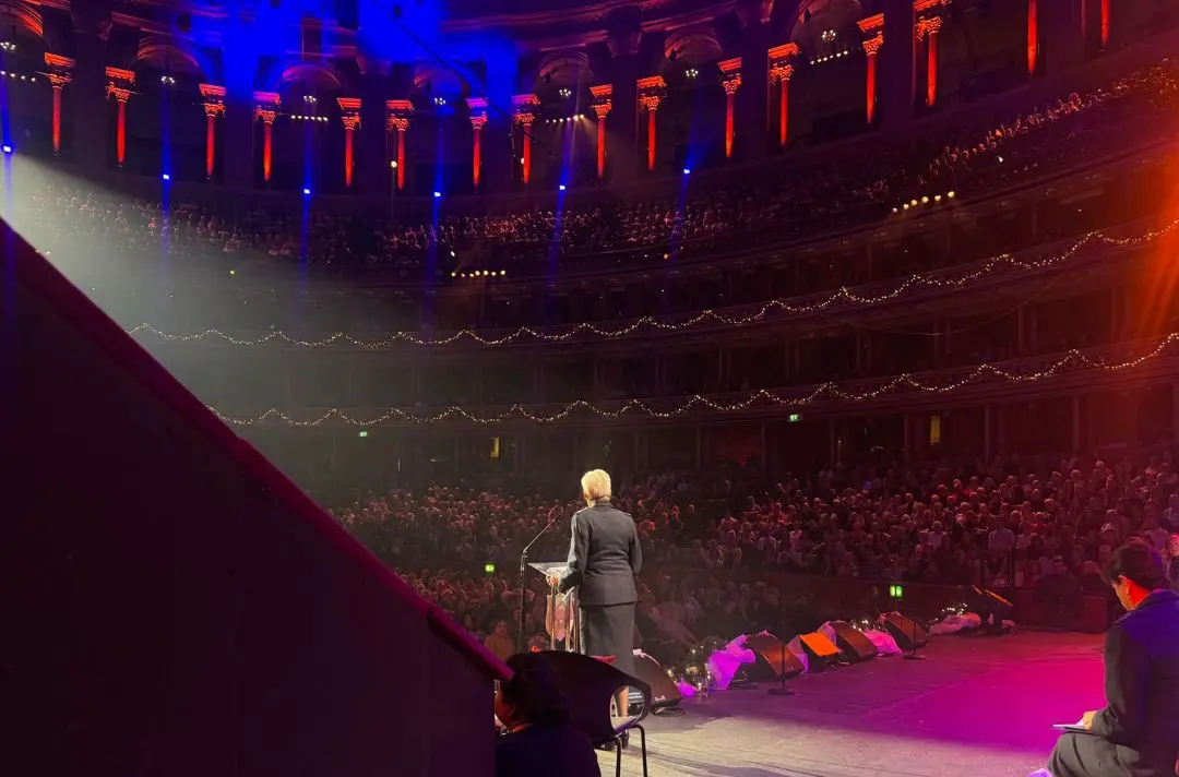 A view from the wings of Jenine Main speaking into a microphone at the lectern on stage at the Royal Albert Hall