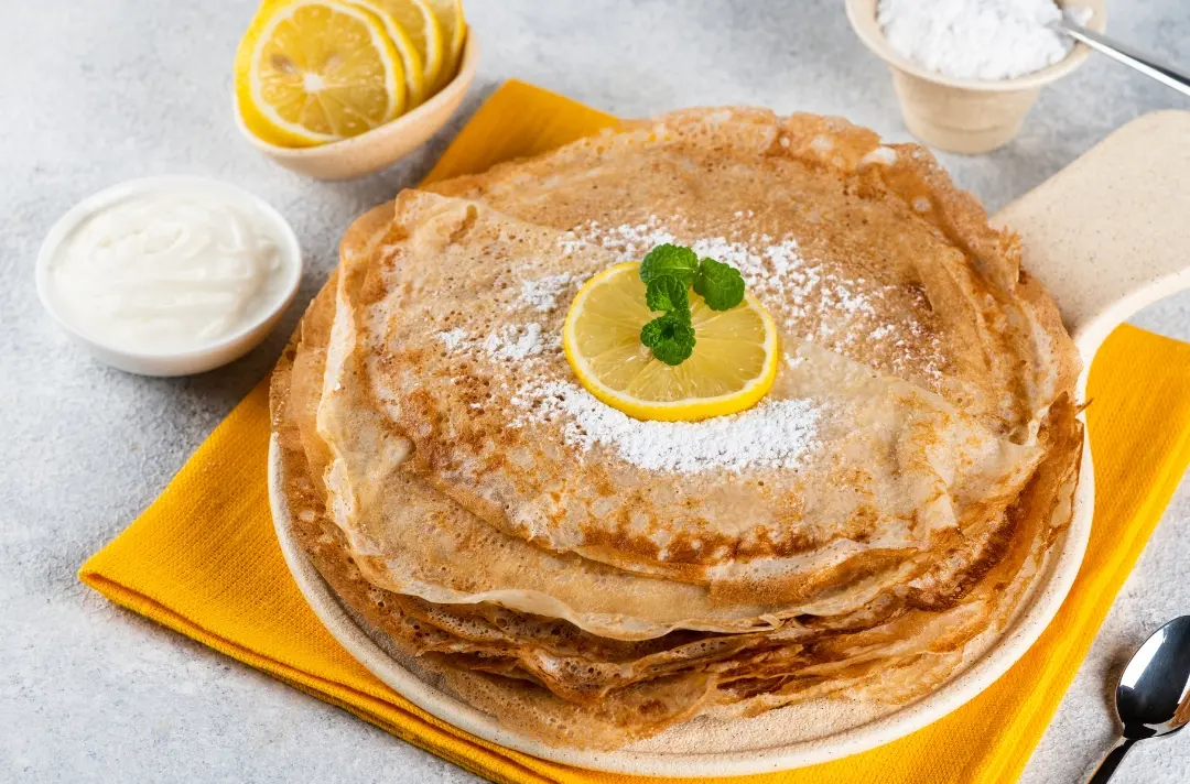A stack of pancakes with lemons around the stack and a slice of lemon on top of the stack