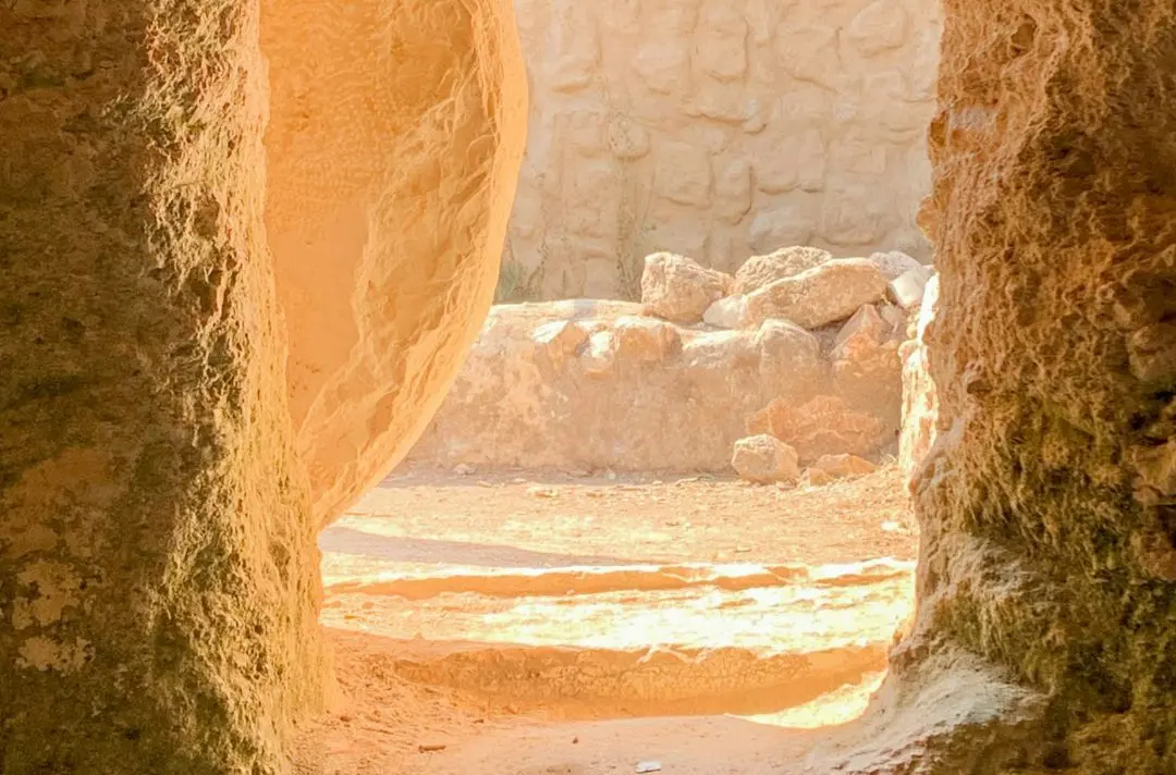 A photo depicting the entrance to a cave with a large stone rolled away from the entrance. The sun is shining down on the entrance.