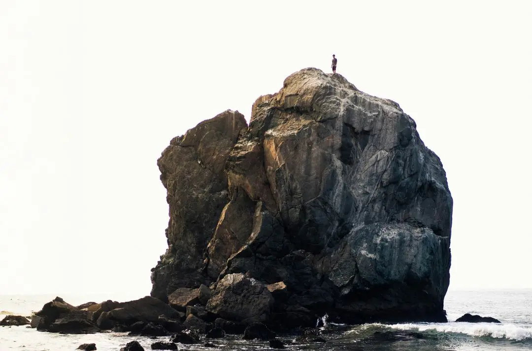A man standing on top of a gigantic rock in the middle of the ocean.
