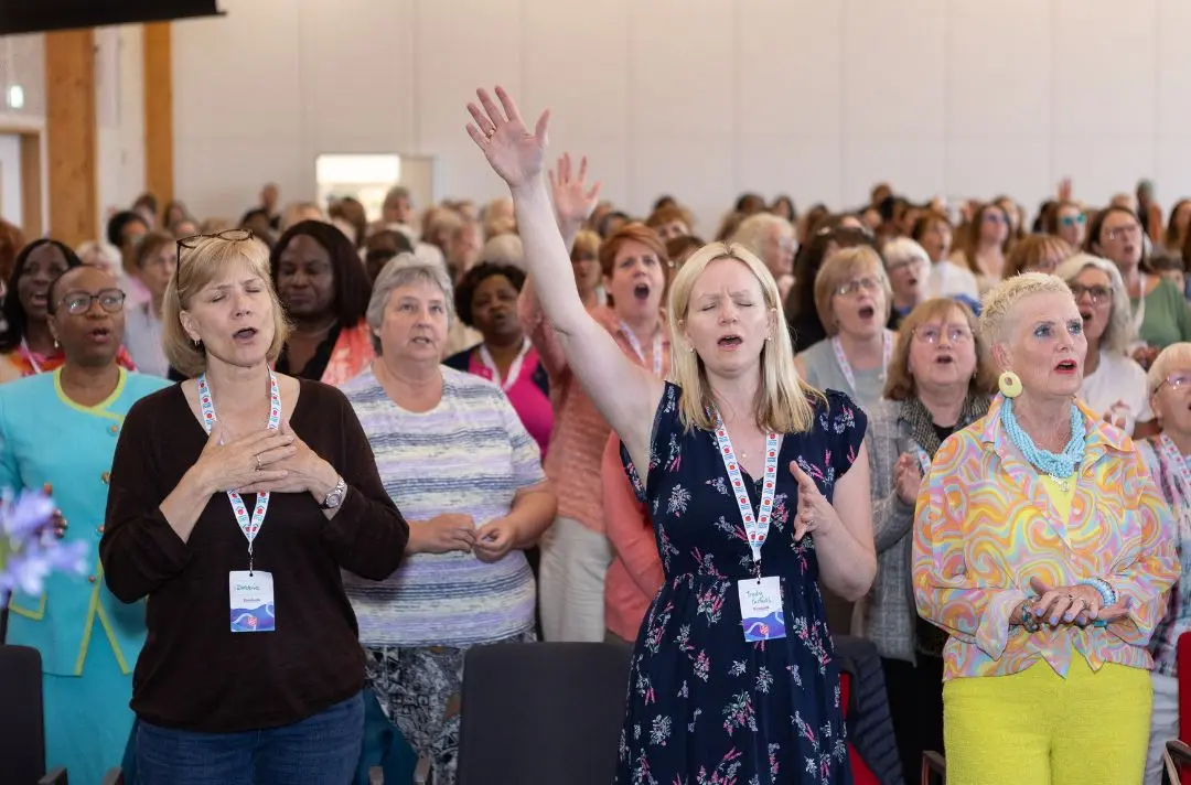 A congregation of women singing. One has her hands up the sky while others close their eyes with their hands to their hearts.
