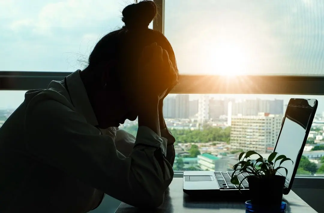 A woman holds her head in her hands despondently while sat a computer desk.