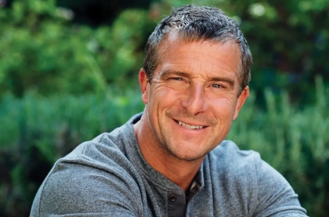 Close-up portrait of an adult person with short grey hair wearing a grey long-sleeve top, sitting outdoors with blurred green foliage in the background. The person is looking toward the camera with a relaxed facial expression, and natural daylight illuminates the face and surrounding greenery.