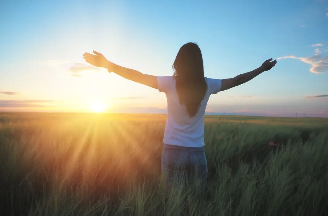 A woman stands in a field of long green grass while holding out her arms facing the rising sun.