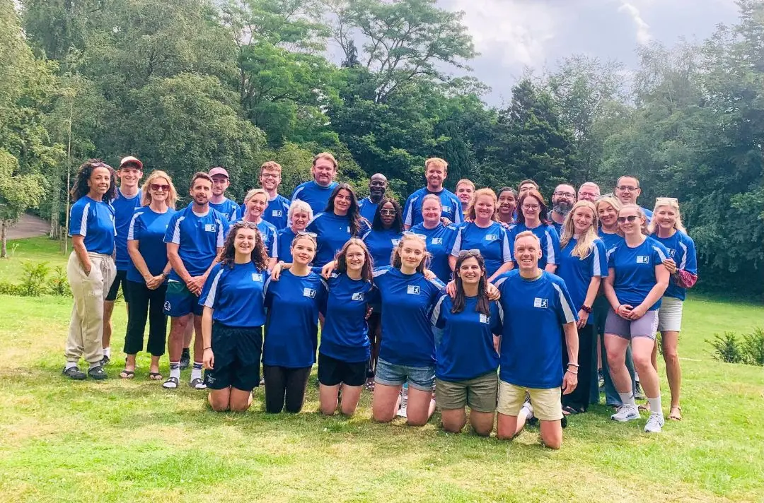 20 to 30 smiling men and women wearing blue sports tops join together for a group photo.
