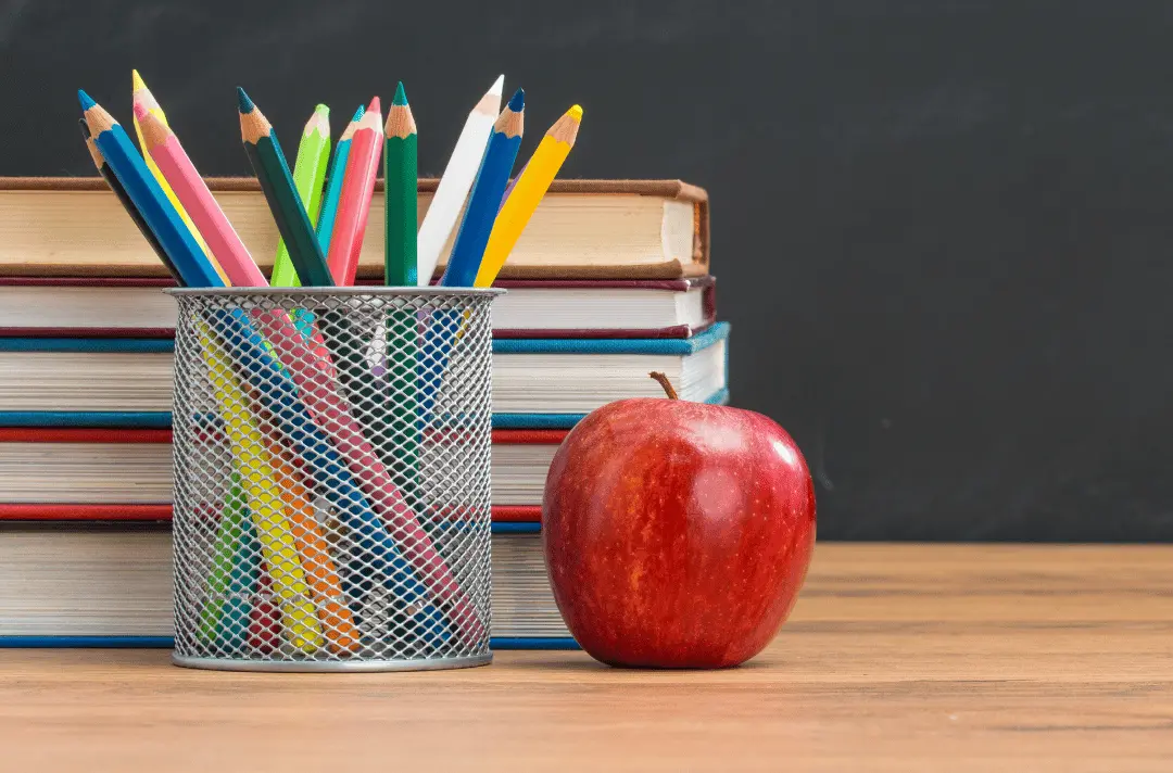 Colourful pencils in a holder, stacked books and a red apple on a classroom desk.