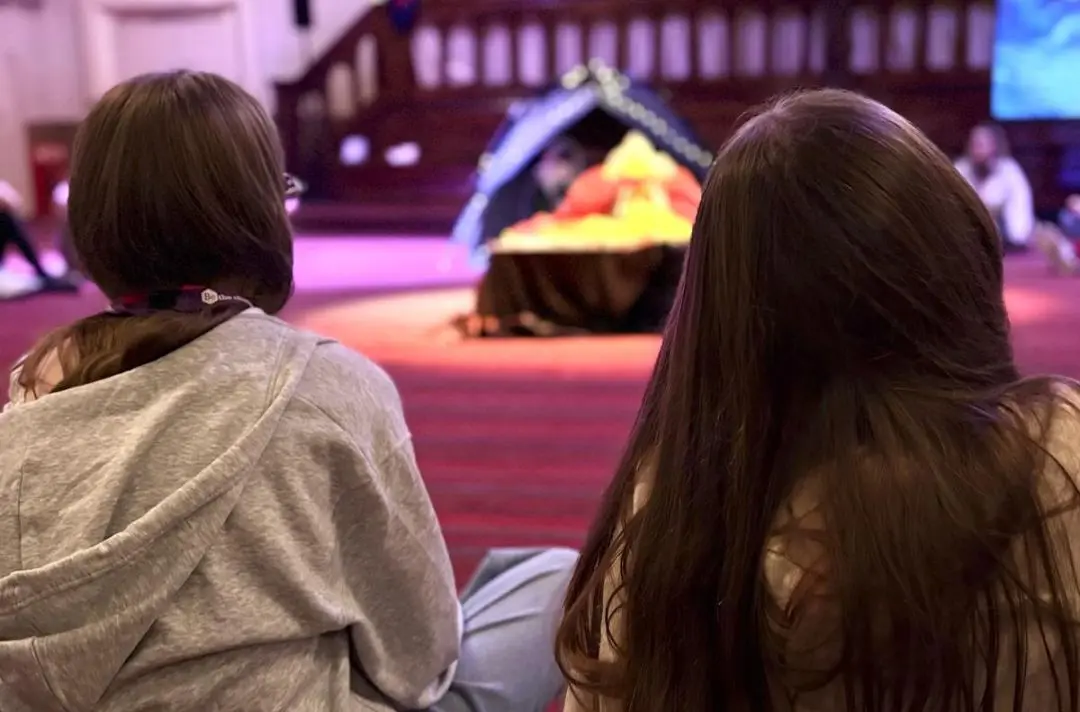 Two girls sit facing a cross.