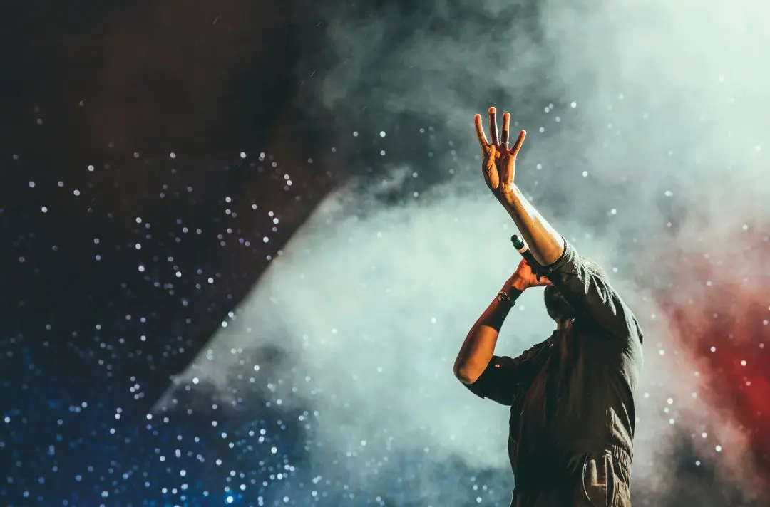 A singer stands with a microphone to his mouth while holding up his hand in the air.