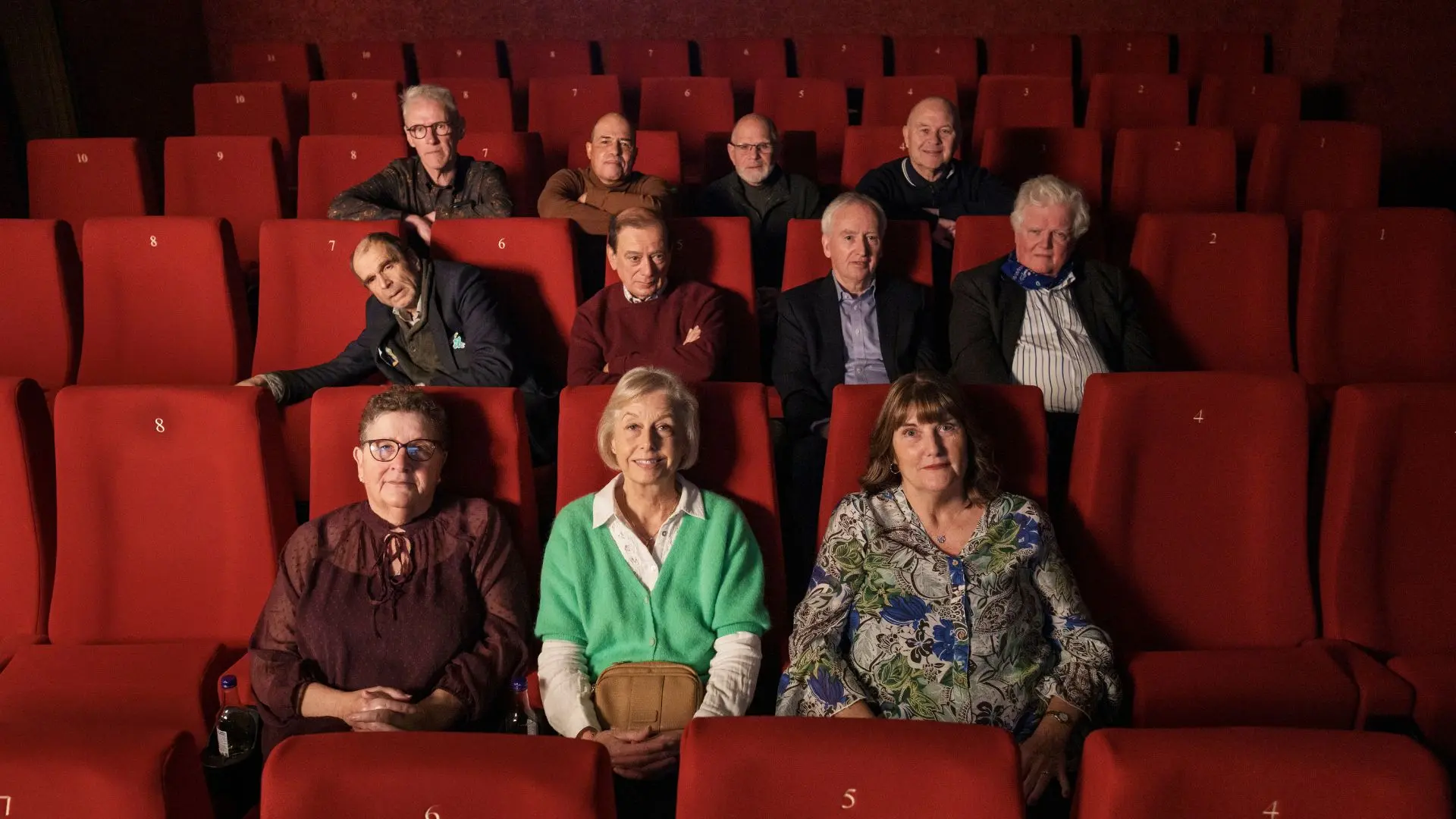 Cast members of 70 Up sit in a cinema facing the camera smiling.