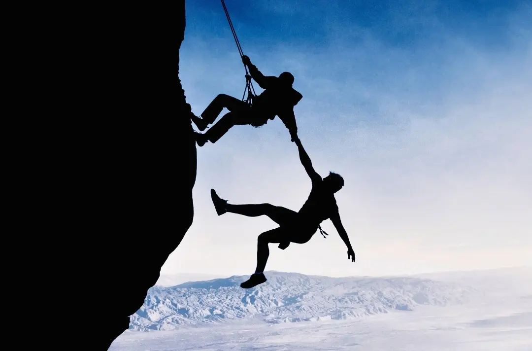 A photo shows two people on a sheer mountainside. One person is secured with a harness, while holding on to the other, who has nothing securing them.