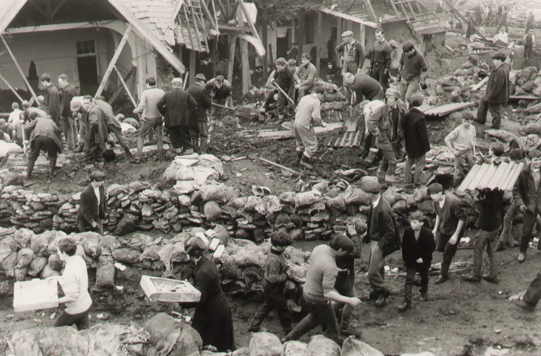 Black-and-white photograph showing dozens of people working together in a muddy disaster area.
