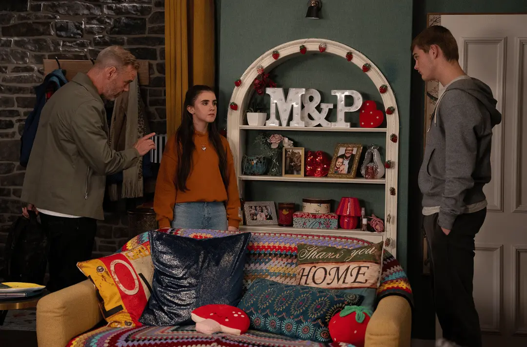 Screen grab from Emmerdale scene of three people standing in a cosy living room. One person on the left gestures with a raised finger while facing two younger people standing apart from each other.