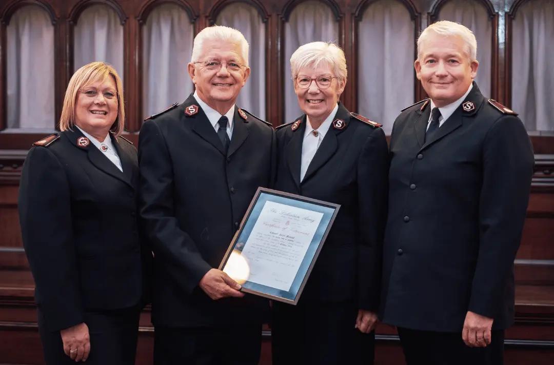 Four Salvation Army officers stand together on a church platform in front of dark wooden panelling. One officer holds a framed Certificate of Retirement while the group poses for a photograph. All are wearing Salvation Army uniforms.