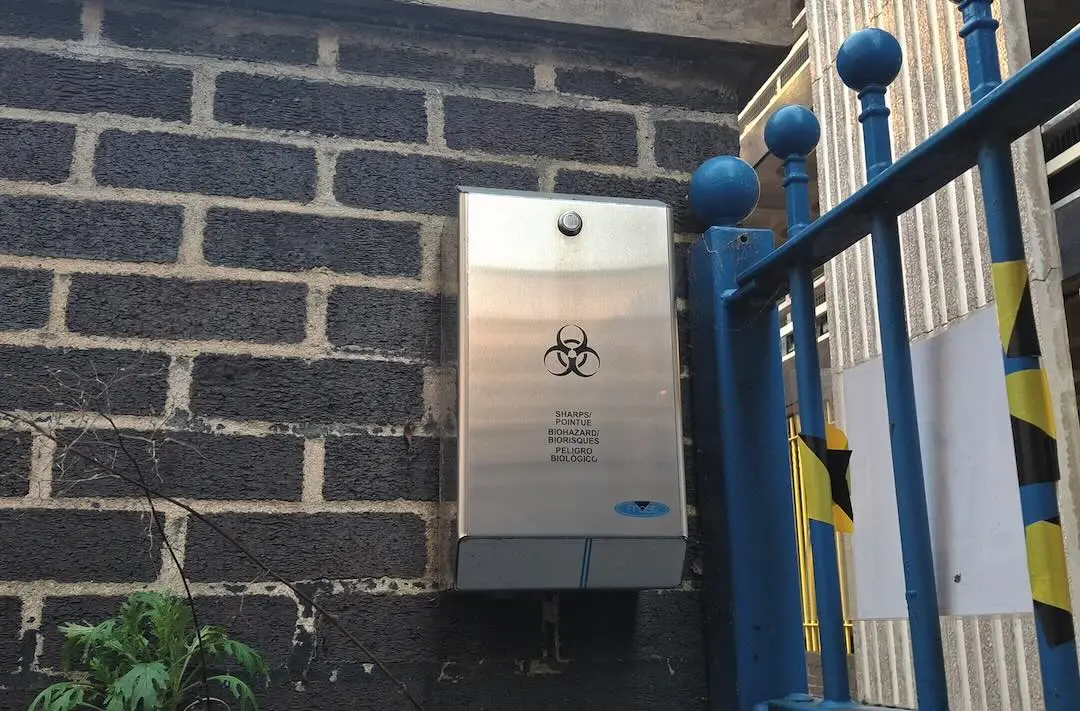 A picture of a metal needle disposal unit against a brick wall outside a Salvation Army Lifehouse.