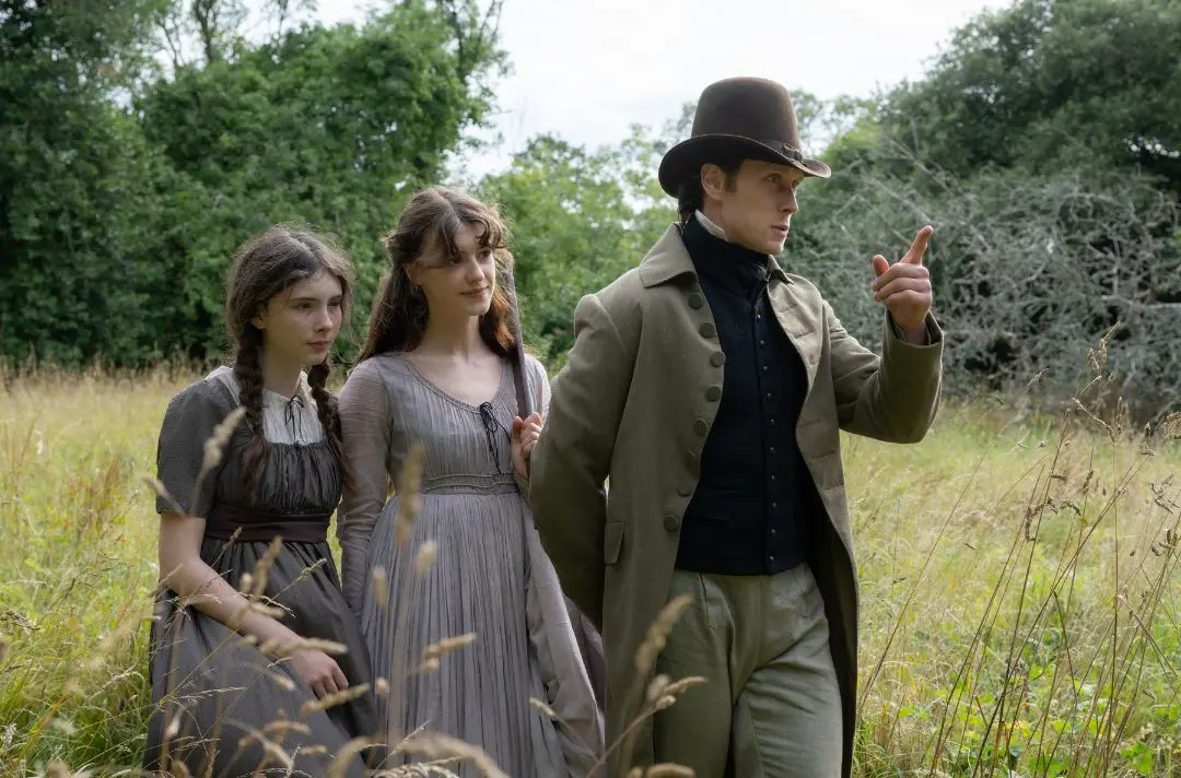 Two young women and a young man, dressed in period costume, take a stroll through a green field. 